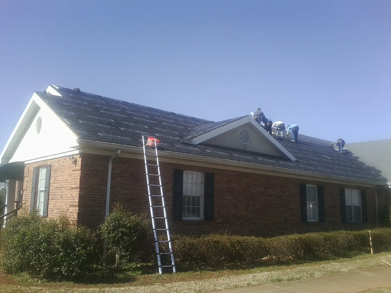 Crew tearing shingles off a brick ranch roof with a ladder set — Emergency Roof Repair work by our crew in South Heart